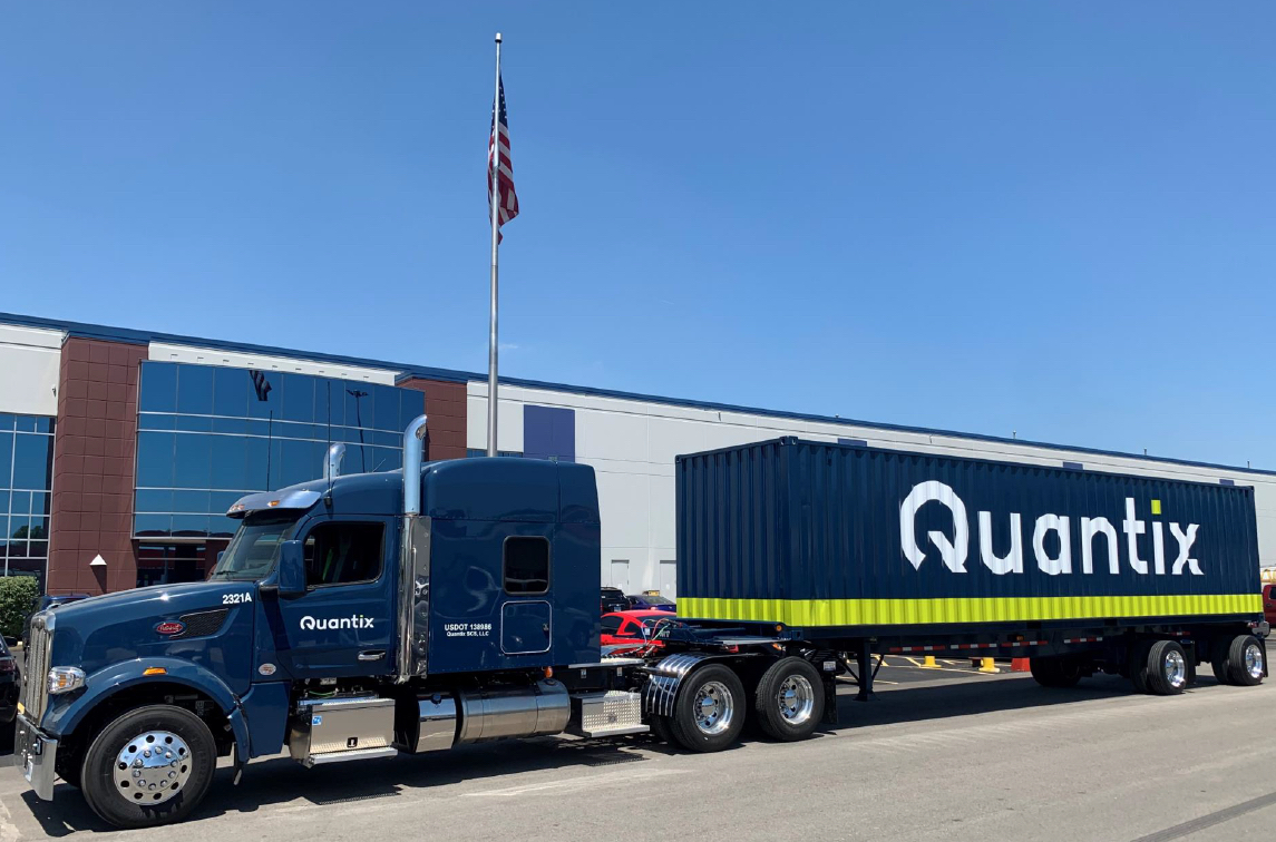 Quantix truck hauls branded container in front of logistics facility under American flag